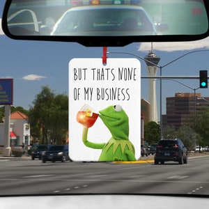 Könnte beinhalten: Ein weißer Auto-Lufterfrischer mit einer grünen Muppet-Figur, die Tee trinkt. Der Text auf dem Lufterfrischer lautet "Aber das geht mich nichts an."