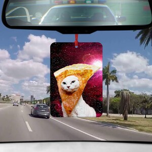 Könnte beinhalten: Eine weiße Katze trägt ein Pizzastück als Hut vor einem Galaxie-Hintergrund. Die Katze schaut in die Kamera. Das Bild ist ein Auto-Lufterfrischer.