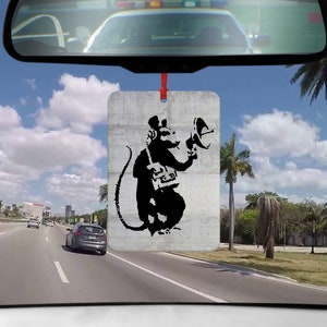 Könnte beinhalten: Ein schwarzer und weißer Auto-Lufterfrischer mit einem Affen, der ein Megaphon hält. Der Affe trägt einen Hut und hat eine Tasche über der Schulter. Der Lufterfrischer hängt an einem Rückspiegel in einem Auto.