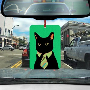 Könnte beinhalten: Ein grüner Auto-Lufterfrischer mit einer schwarzen Katze, die eine Krawatte und ein weißes Hemd trägt. Die Katze schaut nach links. Der Lufterfrischer hängt an einem Rückspiegel.