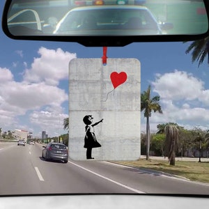Könnte beinhalten: Ein Auto-Lufterfrischer mit einem Schwarzweißbild eines Mädchens, das einen roten herzförmigen Ballon hält. Das Bild ist auf einem grauen Hintergrund mit einem abgenutzten Look.