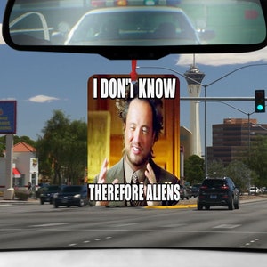 May include: A car air freshener with a meme image of a man with a confused expression. The text on the air freshener reads "I don't know therefore aliens".