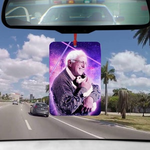 Könnte beinhalten: Ein Auto-Lufterfrischer mit einem Foto eines Mannes, der eine weiße Katze in seinen Armen hält. Das Foto ist vor einem lila und rosa Galaxie-Hintergrund mit einem rosa Laserstrahl platziert.