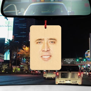 May include: A brown wooden car air freshener with a picture of Nicolas Cage's face on it. The air freshener is hanging from a red string.