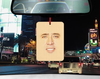 May include: A brown wooden car air freshener with a picture of Nicolas Cage's face on it. The air freshener is hanging from a red string.