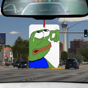 Könnte beinhalten: Ein grüner Comic-Frosch mit einem blauen Hemd wischt sich eine Träne aus dem Auge. Der Frosch schaut nach rechts. Das Bild ist auf weißem Hintergrund.