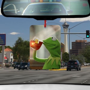 Könnte beinhalten: Eine grüne Froschpuppe mit einer roten Tasse in der Hand, die aus der Tasse trinkt. Der Frosch trägt ein grünes Hemd und hat einen weißen Hintergrund. Das Bild ist ein Auto-Lufterfrischer.