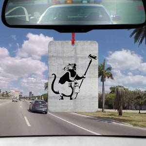 Könnte beinhalten: Ein Auto-Lufterfrischer mit einer schwarz-weißen Schablone einer Ratte, die eine Farbrolle hält. Die Ratte befindet sich auf einem grauen Betonuntergrund.