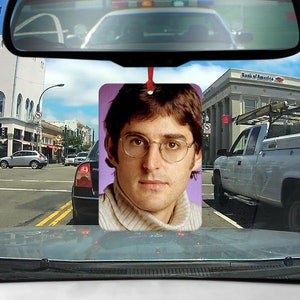 Könnte beinhalten: Ein Auto-Lufterfrischer mit einem Porträt eines Mannes mit braunen Haaren und Brille, der einen hellbraunen Pullover trägt. Der Hintergrund ist helllila.