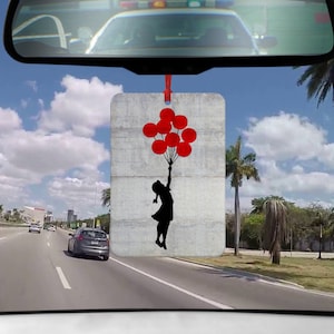 Könnte beinhalten: Rechteckiger Auto-Lufterfrischer mit einer Silhouette eines Mädchens, das rote Ballons vor einer grauen Betonmauer hält. Der Lufterfrischer hängt an einem roten Band, mit Blick auf eine Autobahn und Palmen im Hintergrund.