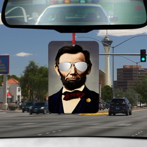 May include: A car air freshener featuring a portrait of Abraham Lincoln wearing sunglasses. The air freshener is a rectangular piece of cardboard with a black background and a white border.
