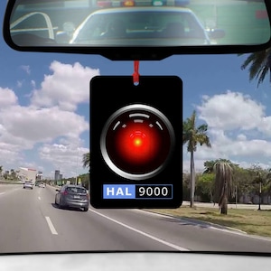 May include: A black car air freshener with a red and silver HAL 9000 logo. The logo features a red eye with a black circle around it. The text "HAL 9000" is in blue letters below the eye.