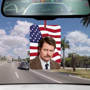 Könnte beinhalten: Ein Auto-Lufterfrischer mit einem Porträt von Ron Swanson aus der Fernsehserie Parks and Recreation. Das Bild ist vor dem Hintergrund der amerikanischen Flagge.