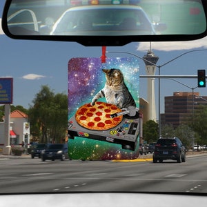 Könnte beinhalten: Ein Auto-Lufterfrischer mit einer Katze, die eine Baseballkappe verkehrt herum trägt und eine Pizza auf einem Plattenspieler hält. Der Hintergrund ist eine Galaxie mit Sternen und einem grün-blauen Nebel.