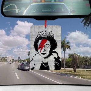 Könnte beinhalten: Ein Auto-Lufterfrischer mit einem Schwarzweißbild von Queen Elizabeth II mit einem roten und blauen Blitz über ihrem Auge. Der Hintergrund ist eine graue Betontextur.