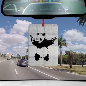 Könnte beinhalten: Rechteckiger Auto-Lufterfrischer mit einem Panda-Bären-Design im Banksy-Stil. Der schwarz-weiße Panda hält zwei Pistolen vor einem grauen Betonhintergrund. Er hängt an einem roten Band, sichtbar durch die Windschutzscheibe eines Autos.