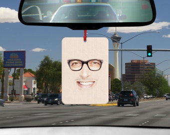 May include: A white car air freshener with a photo of a man wearing glasses and smiling. The air freshener is hanging from a rearview mirror in a car.