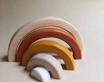May include: A set of five wooden rainbow arches in natural wood tones, including beige, yellow, orange, and brown. The arches are stacked on top of each other, creating a rainbow shape.