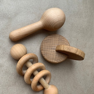 May include: A collection of three wooden baby toys. The toys include a maraca-style rattle, a circular rattle with interlocking rings, and a small wooden disc with a handle. All items are made of light-colored wood.