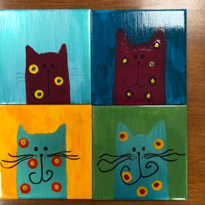 May include: Four square paintings of cats with different coloured backgrounds. The cats are painted in a whimsical style with large eyes and a simple design. The backgrounds are blue, green, yellow, and teal.