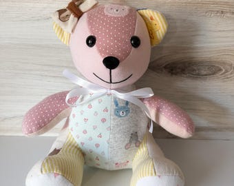 May include: A stuffed bear toy made from various patterned fabrics in shades of pink, yellow, and light blue. The bear features a white ribbon tied around its neck, a small bow on its left ear, and the name Iris embroidered on its left foot.