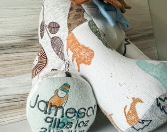May include: A stuffed animal toy made from various patterned fabrics featuring lions and leaves. The foot pad is embroidered with the name Jameson, the weight 9 lbs 1 oz, and the length 21 3/4 in. It has a brown fabric mane and a blue bow tie.