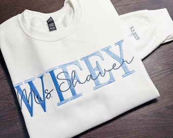 May include: Cream-colored sweatshirt with the word "WIFEY" in blue block letters and "Mrs. Shaver" in a cursive font. The sleeve displays the date "5.3.2025" in blue. A casual, comfortable garment.