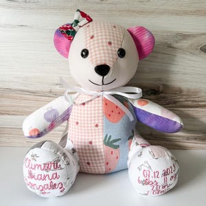May include: A patchwork teddy bear featuring a light pink gingham pattern with small strawberries, a blue section with large strawberries, and purple accents. It has a white ribbon bow, black button eyes, and embroidered text on its feet.