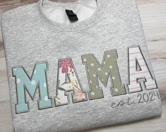 May include: A light gray crewneck sweatshirt featuring the word MAMA in large block letters. Each letter has a unique pattern: light blue with white dots, floral print, sage green with white flowers, and pink stripes. The text est. 2024 is embroidered below.