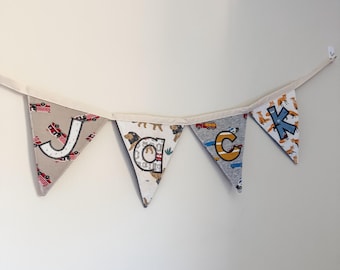 May include: A fabric bunting banner spelling the name JACK with four triangular pennants. Each pennant features a unique pattern including fire trucks, lions, cars, and foxes, with large appliqued letters attached to the front.