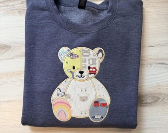 May include: A dark heather gray crewneck sweatshirt featuring a large, central teddy bear appliqué. The bear is constructed from various patterned fabric patches, including images of sheep, fire trucks, rainbows, and butterflies.
