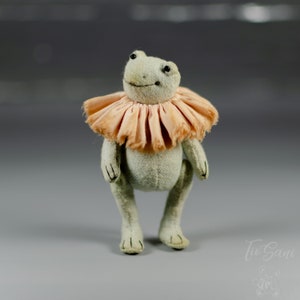 May include: A handmade frog doll with a light green body and a peach-colored ruffled collar. The frog has black bead eyes and stitched details. The doll is standing on its hind legs.
