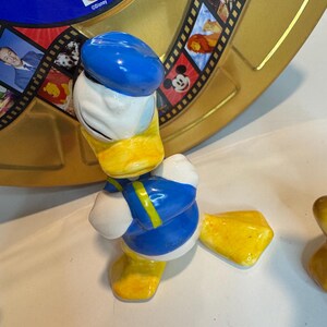 May include: A ceramic figurine of Donald Duck wearing a blue sailor hat and a blue and yellow sailor shirt. He is standing with his arms crossed and has a yellow beak and feet.