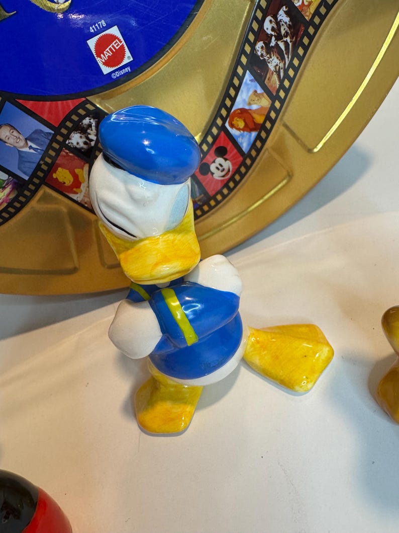 May include: A ceramic figurine of Donald Duck wearing a blue sailor hat and a blue and yellow sailor shirt. He is standing with his arms crossed and has a yellow beak and feet.