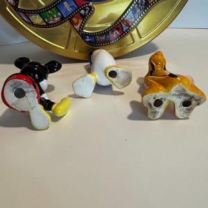 May include: Three ceramic figurines of Disney characters: Mickey Mouse, Donald Duck, and Pluto. Mickey Mouse is black with red shorts and yellow shoes. Donald Duck is yellow with a blue sailor shirt and white pants. Pluto is brown with a yellow collar. The figurines are all standing on a white surface.