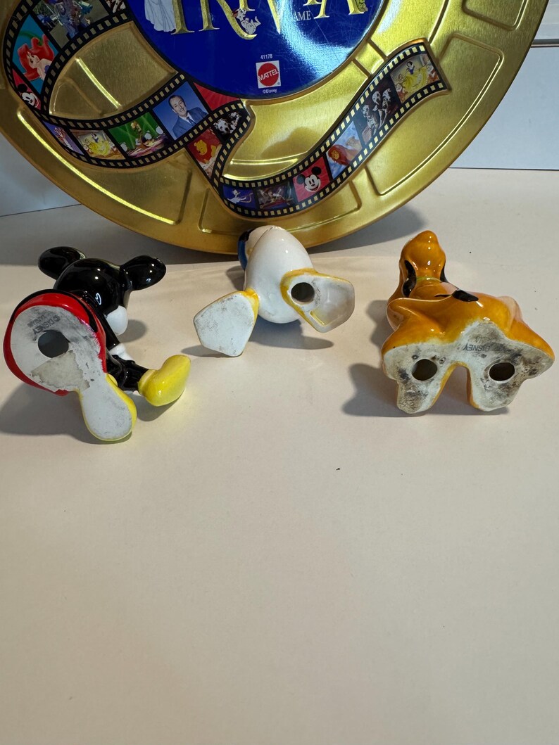 May include: Three ceramic figurines of Disney characters: Mickey Mouse, Donald Duck, and Pluto. Mickey Mouse is black with red shorts and yellow shoes. Donald Duck is yellow with a blue sailor shirt and white pants. Pluto is brown with a yellow collar. The figurines are all standing on a white surface.