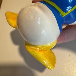 May include: A ceramic figurine of a cartoon duck wearing a blue and yellow shirt. The duck is standing on its feet with its head tilted to the side. The figurine is painted in bright colors and has a glossy finish.