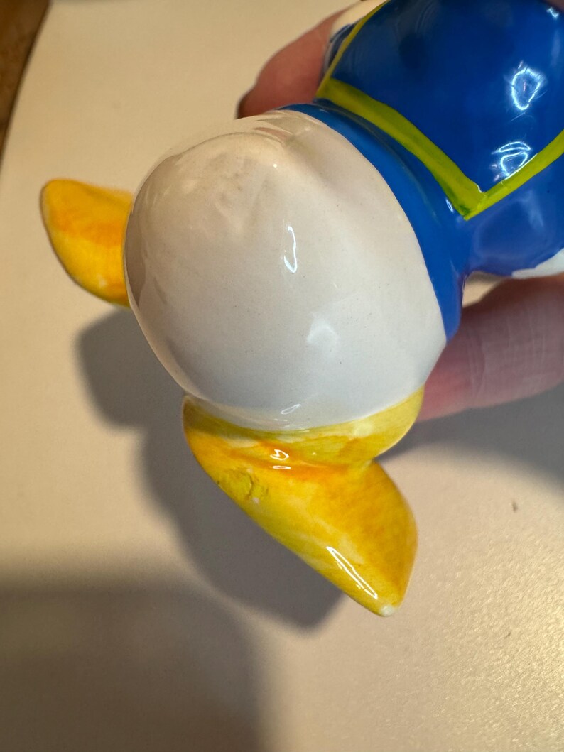 May include: A ceramic figurine of a cartoon duck wearing a blue and yellow shirt. The duck is standing on its feet with its head tilted to the side. The figurine is painted in bright colors and has a glossy finish.