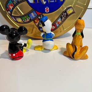 May include: Three ceramic figurines of Disney characters: Mickey Mouse in red shorts with white polka dots, Donald Duck in a blue sailor suit, and Pluto in orange with a black collar. The figurines are sitting on a white surface in front of a gold and blue game board with the word "Trivia" printed on it.