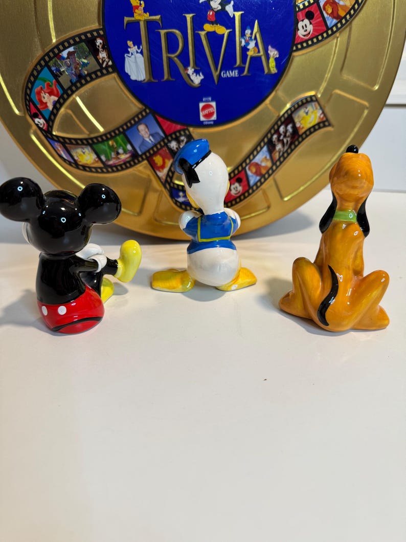 May include: Three ceramic figurines of Disney characters: Mickey Mouse in red shorts with white polka dots, Donald Duck in a blue sailor suit, and Pluto in orange with a black collar. The figurines are sitting on a white surface in front of a gold and blue game board with the word "Trivia" printed on it.