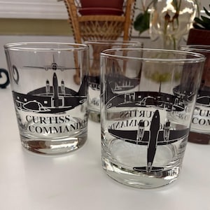 May include: Set of four clear drinking glasses featuring a black Curtiss Commando airplane design. The glasses have a lowball shape and the text "Curtiss Commando" is printed on the front. The glasses are suitable for serving beverages.
