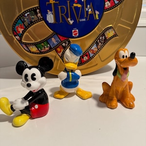 May include: Three ceramic figurines of Disney characters: Mickey Mouse, Donald Duck, and Pluto. Mickey Mouse is wearing his signature red shorts and yellow shoes. Donald Duck is wearing a blue sailor shirt and yellow pants. Pluto is wearing a brown and orange dog costume. The figurines are sitting on a white surface.