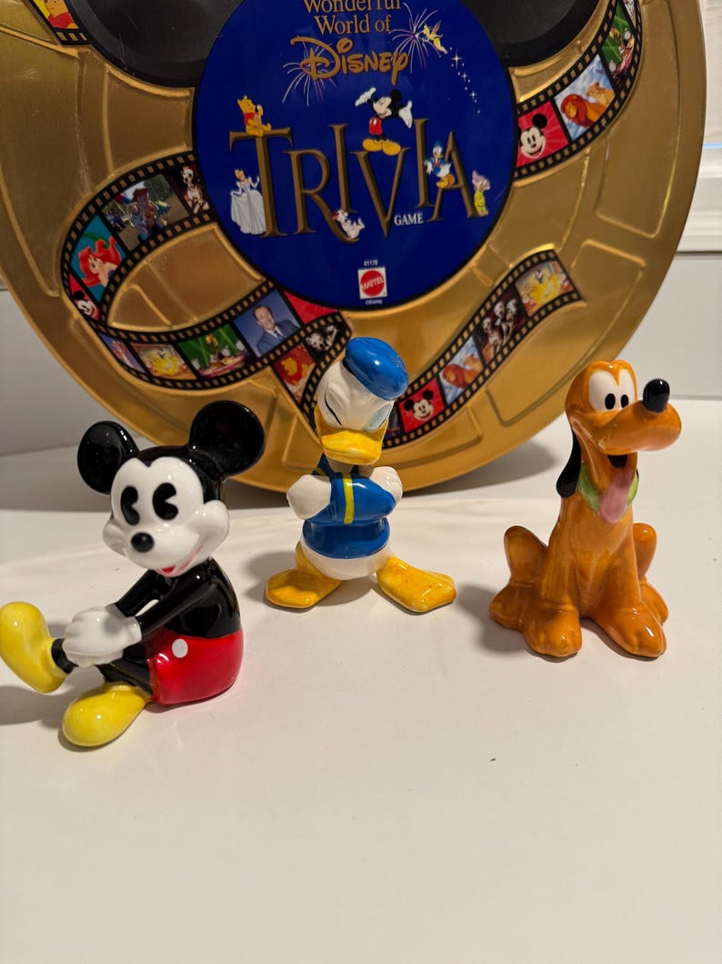 May include: Three ceramic figurines of Disney characters: Mickey Mouse, Donald Duck, and Pluto. Mickey Mouse is wearing his signature red shorts and yellow shoes. Donald Duck is wearing a blue sailor shirt and yellow pants. Pluto is wearing a brown and orange dog costume. The figurines are sitting on a white surface.