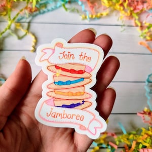Op de afbeelding: Een sticker met een stapel strohoeden met kleurrijke linten. De sticker heeft de tekst "Join the Jamboree" in een bannerontwerp. De sticker wordt in een hand gehouden tegen een achtergrond van kleurrijke papiersnippers.