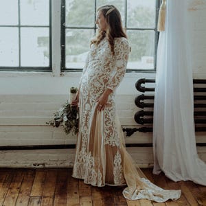 May include: A woman in a white lace wedding dress with a long train stands in front of a window. She is holding a bouquet of flowers in her left hand.