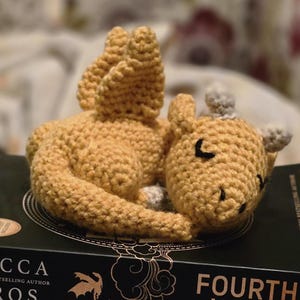 Puede incluir: Una pila de tres libros de Rebecca Yarros, con los títulos "Fourth Wing", "Iron Flame" y "Onyx Storm". Una figura de dragón amarillo tejido a ganchillo descansa encima de los libros. Los libros son negros con letras doradas.