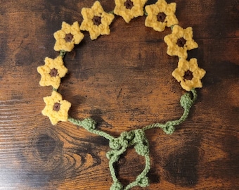 May include: A crocheted sunflower headband with green leaves. The headband is made of yellow and brown yarn and is tied with a bow at the back.