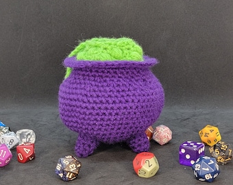 Può includere: Un calderone di uncinetto viola con un coperchio di uncinetto verde. Il calderone è seduto su una superficie nera e circondato da dadi colorati.
