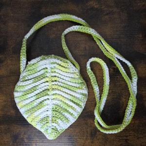 May include: A crocheted leaf-shaped bag with a green and white striped pattern. The bag has a long strap that can be worn over the shoulder.