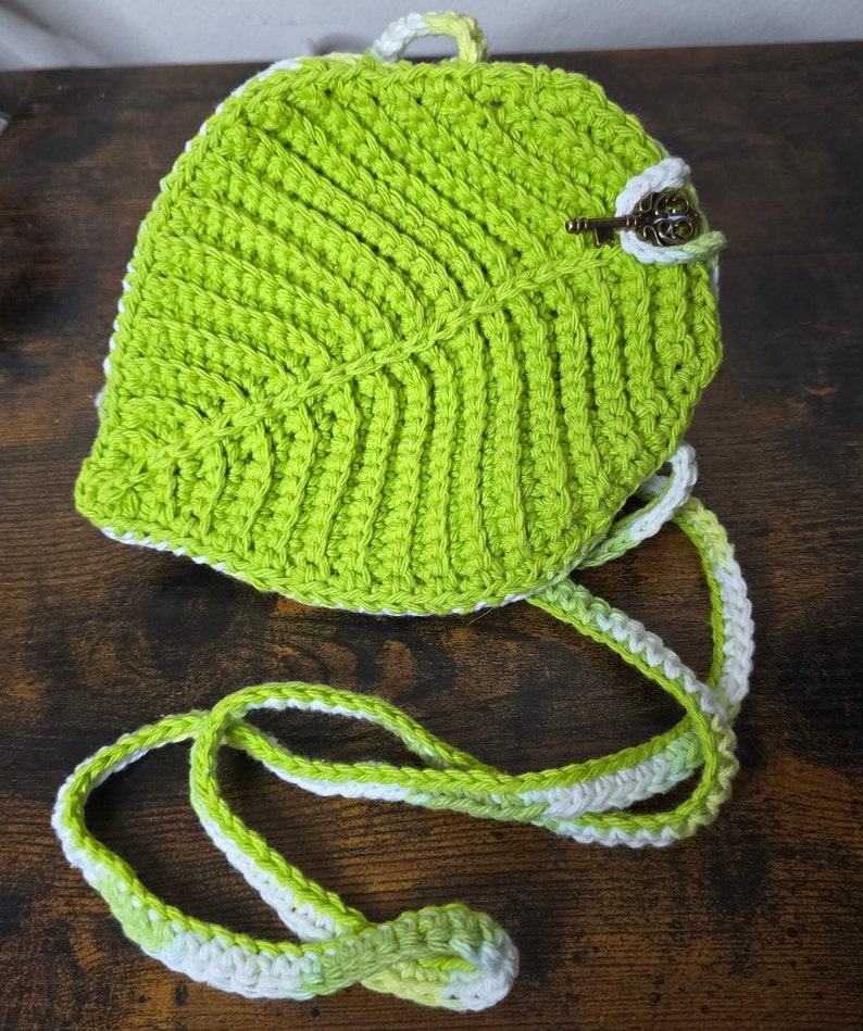 May include: A green crocheted leaf-shaped bag with a white and green crocheted strap. The bag has a small key charm attached to it.